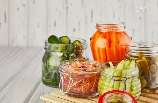 Glass jars of fermented vegetables (cucumbers, carrots, celery) on a wooden surface — 發酵食物對身體的益處 · The benefits of fermented foods